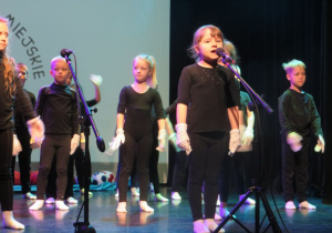 dziewczynka śpiewa do mikrofonu, dzieci w tle prezentują układ taneczny