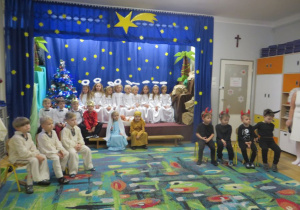 Dzieci z grupy III siedzą na scenie podczas jasełkowego przedstawienia przebrane za anioły, diabełki, królów, pasterzy, Maryję i Józefa