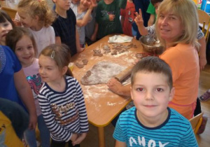 Dzieci z grupy IV stoją przy stole i patrzą jak Pani wałkuje ciasto na pierniki