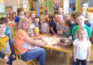 Dzieci z grupy IV stoją przy stole i patrzą jak Pani wałkuje ciasto na pierniki