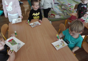 Troje dzieci nakleja na papierowych talerzykach obrazki produktów mlecznych wyciętych z gazetek sklepowych