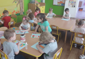 Dzieci z grupy I siedzą przy stolikach i naklejają na kartkach figury geometryczne na kształt cxzłonków swojej rodziny