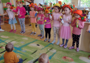 8 dziewczynek ma kapelusze ozdobione owocami i warzywami, grają grzechotkami wykonanymi z pojemników po jogurcie