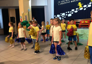 Dzieci przebrane w żółte koszulki i niebieskie spodnie tańczą z pomponami do muzyki z bajki o Minionkach