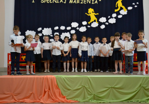 Dzieci stoją na scenie, jedna dziewczynka do mikrofonu czyta tekst z kartki
