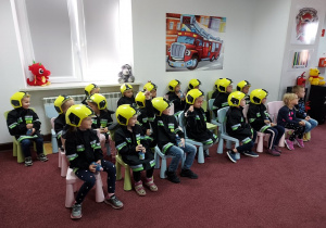 Dzieci siedzą na krzesełkach ubrane w mundur strażacki i hełm