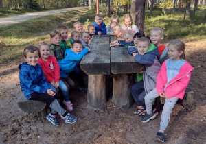 Dzieci z grupy V odpoczywają siedząc na lesnej ławce ze stoliczkiem