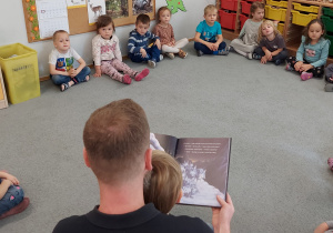 Dzieci siedzą w kole słuchając bajki czytanej przez tatę Tytusa