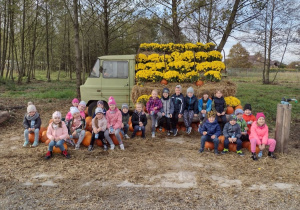 Przedszkolaki z grupy V siedzą przed samochodem przybranym dyniami i żółtymi chryzantemami