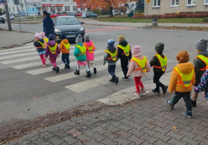 Dzieci z grupy II ubrane w kamizelki odblaskowe przechodzą przez skrzyżowanie po pasach