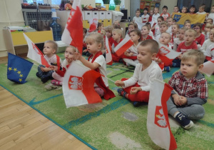 Młodsze przedszkolaki siedzą na dywanie i trzymają małe flagi Polski