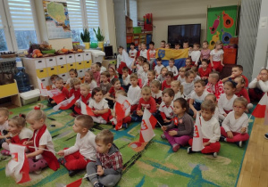 Przedszkolaki siedzą na dywanie w biało-czerownych strojach podczas śpiewania piosenki o symbolach narodowych
