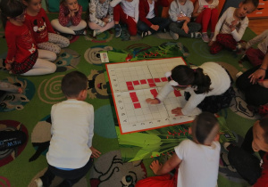 Dzieci siedzą na dywanie i obserwują jak dziewczynka układa kwadrat na macie według kodu