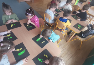 Dzieci za pomocą słomki rozdmuchują zieloną i żółtą farbę na czarnej kartce