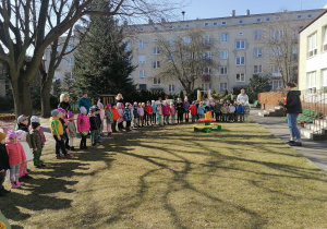 Przedszkolaki stoją w ogrodzie ustawione grupami i słuchają co prowadząca przygotowała dla nich z okazji pierwszego dnia wiosny