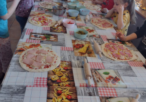 Przedszkolaki stoją przy długim stole i nakładają wybrane składniki na pizze