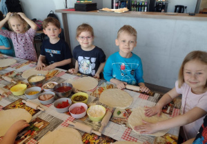Pięcioro dzieci stoi przy swoich rozwałkowanych ciastach na pizze