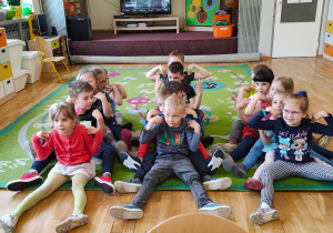 Dzieci siedzą na dywanie w trzech rzędach podczas pląsu "Gąsienica Basi"