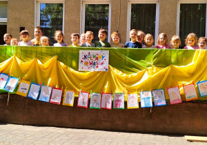 Przedszkolaki z grupy IV stoją przy dekoracji w ogrodzie, na której są portrety rodziców narysowane przez dzieci