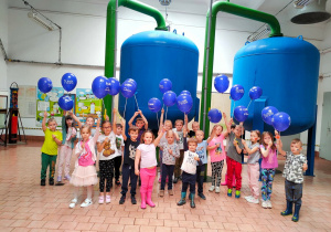Przedszkolaki stoją w stacji uzdatnia wody z niebieskimi balonami