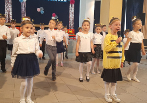 Dzieci stoją w rozsypce i tańczą do piosenki o pszczółce, jedna dziewczynka ma strój pszczółki i śpiewa piosenkę do mikrofonu