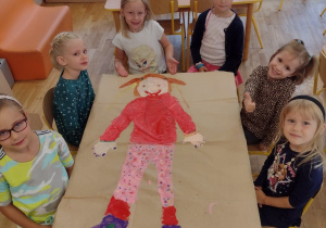 Dziewczynki z grupy piątej siedzą wokół obrazka dziewczynki z przedszkola namalowanego przez siebie
