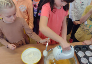 Dziewczynka miesza ciasto mikserem, a druga dosypuje jej maki do miski