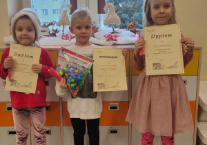 Trzy dziewczynki stoją i pokazują dyplomy otrzymane za udział w konkursie plastycznym
