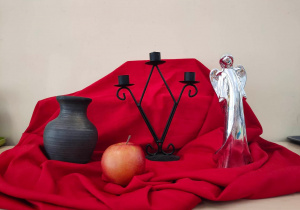 Na czerwonym materiale ustawiony jest wazon, świecznik, jabłko i anioł