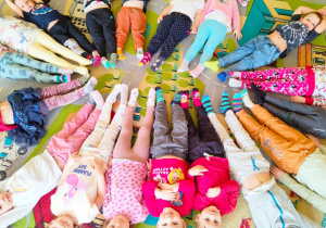 Przedszkolaki z grupy pierwszej leżą na dywanie i pokazują swoje kolorowe skarpetki