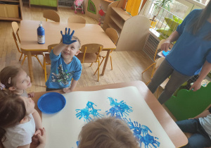 Dzieci z grupy trzeciej maczają swoje ręce w niebieskiej farbie i odbijają je na karonie