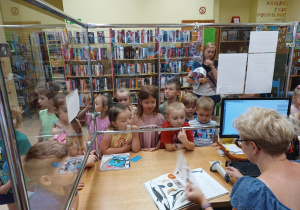 Dzieci przypatrują się pracy pani bibliotekarki podczas wypożyczania przez kogoś książki