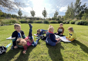 Dzieci wykonują zadania na kartkach siedząc na trawie w Afloparku