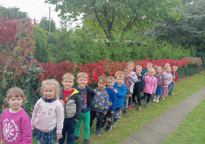 Przedszkolaki stoją jeden za drugim przy płocie oplecionym czerwonymi liśćmi