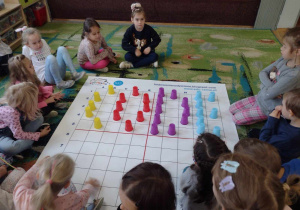 Dzieci siedzą wokół maty z napisem z kolorowych kubeczków code