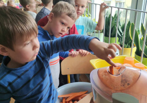 Przedszkolaki stoją w kolejne, aby włożyć marchewki do sokownika