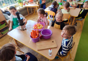 Dzieci z grupy IV siedzą przy stoliku, na którym jest miska z marchewką i sokownik