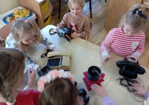 Dziewczynki oglądają przy stoliku stare telefony, aparat oraz młynek