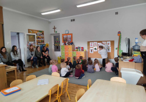 Dzieci siedzą na dywanie przed tablicą i słuchają nauczycielki