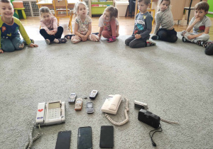 Dzieci siedzą w kole i patrzą na stare aparaty oraz telefony