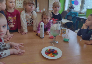 Przedszkolaki stoją przy stolikach i obserwują kolorową wodę