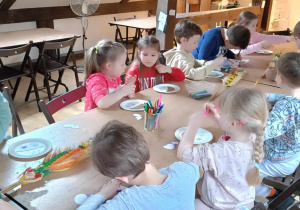 Dzieci siedzą przy stolikach i robią koszycki z talerzyków papierowych