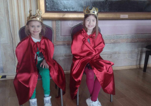 Dwie dziewczynki siedzą na tronach w czerwonych pelerynkach i z koroną na głowie