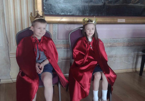 Dwoje dzieci siedzi na tronach w czerwonych pelerynkach i z koroną na głowie