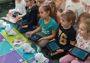 Dzieci siedzą na dywanie i słuchają o robotach, które stoją przed nimi